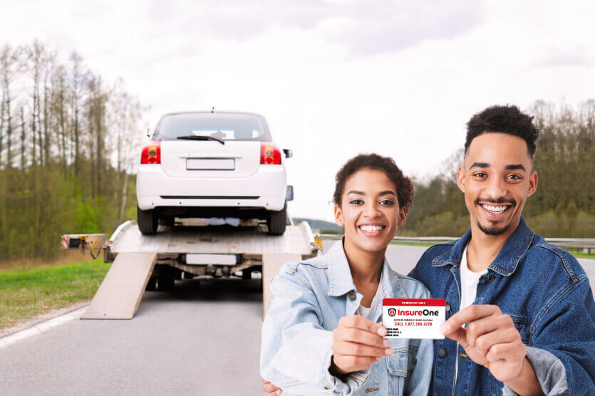 Smiling couple showing Insure One Road Assistance card as their car in the background is being towed away on the highway