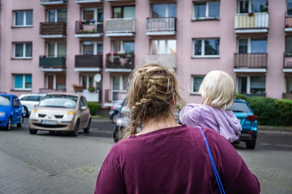 A woman carrying her child seen from behind to their apartments with cars parked in front.