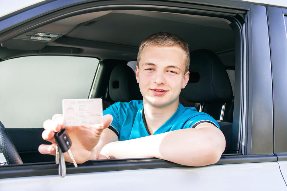 Smiling teenage boy behind the wheel shows his driver's license and holds the keys - best car insurance for teen drivers.