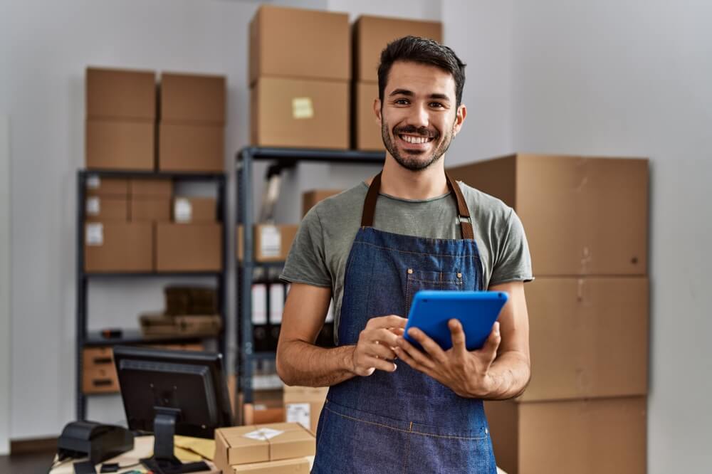 Young Hispanic worker in small warehouse smiling, happy to have workers comp benefits.