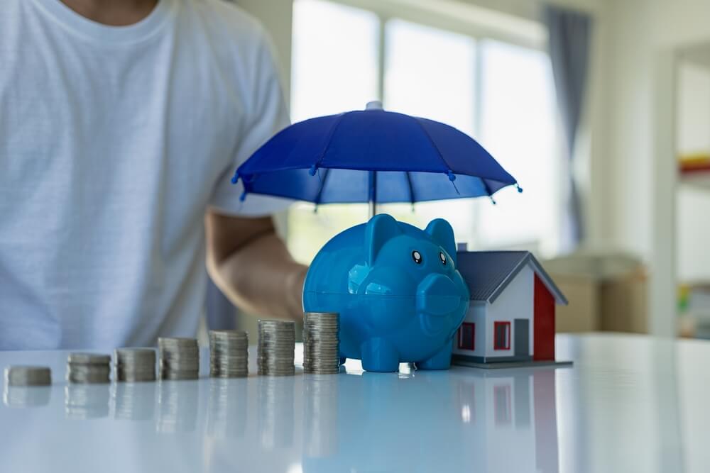 Man stands next to an image of home hazard protection with a house, piggybank and coins under an umbrella - best homeowners insurance.