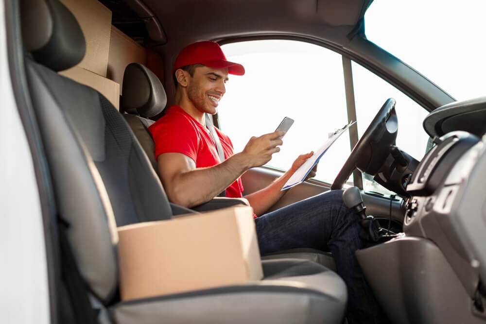 Delivery driver sitting inside a van checking orders on his phone and clipboard, highlighting the need for delivery driver car insurance while on the road.
