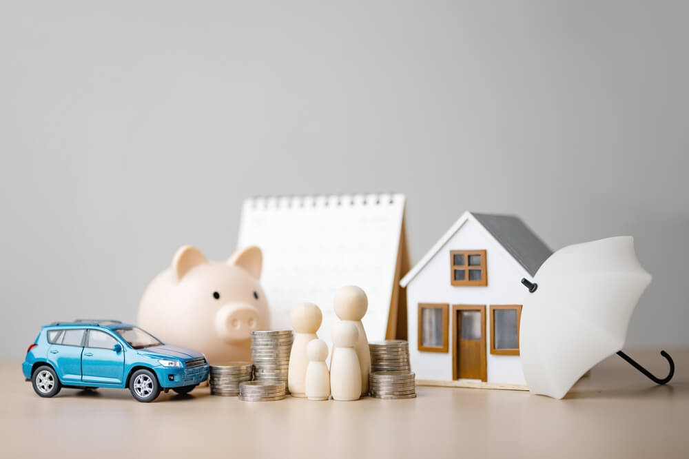 Miniature house, car, family figures, coins, and an umbrella arranged together, visually explaining the concept of insurance bundling and combined financial protection.