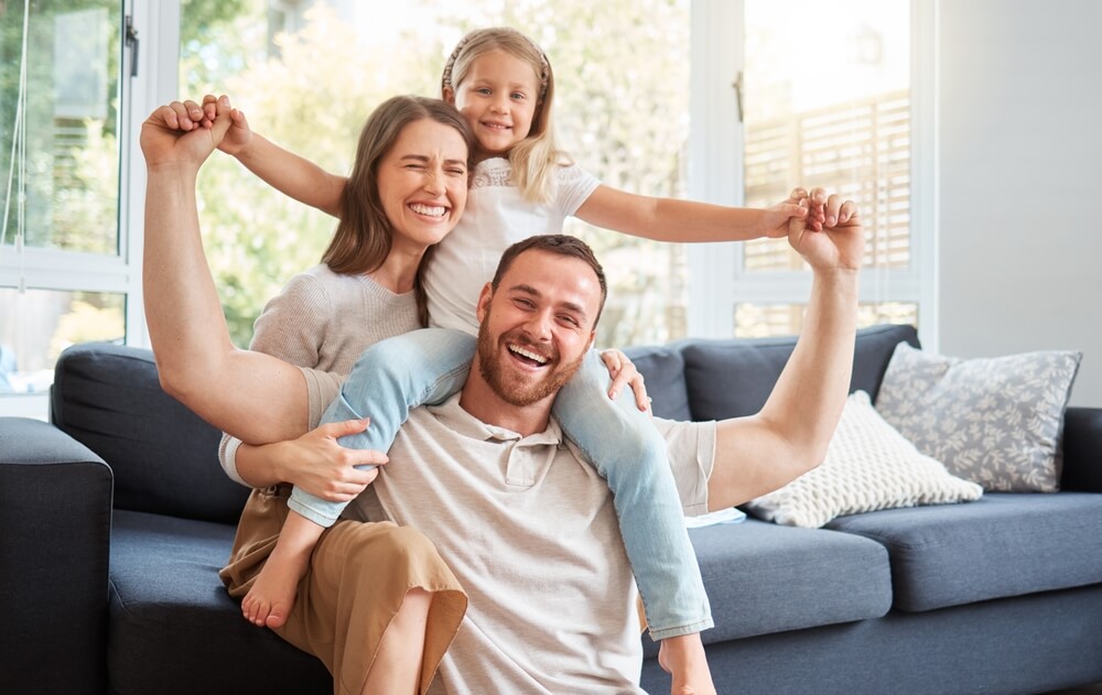 Smiling family in their living room, dad sits on floor with little girl sitting on his neck and mom sits on couch and grinning.