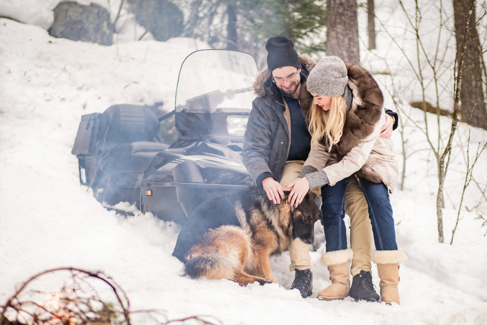 Woman and man with dog on snowy trail, snowmobile in the background