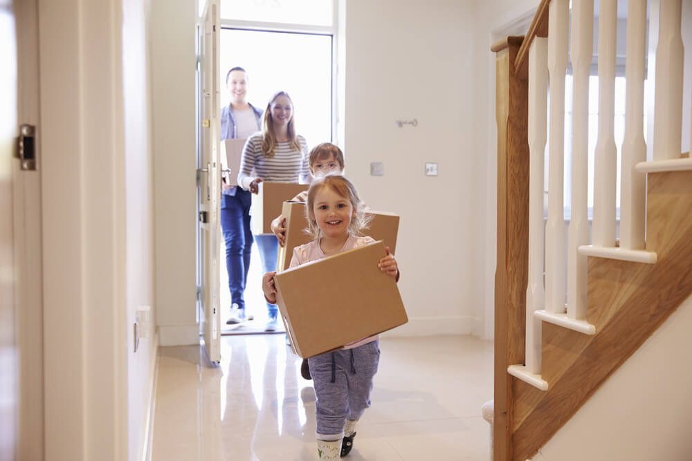 Family arriving at a new home because their house is being repaired, carrying cardboard boxes with their belongings.
