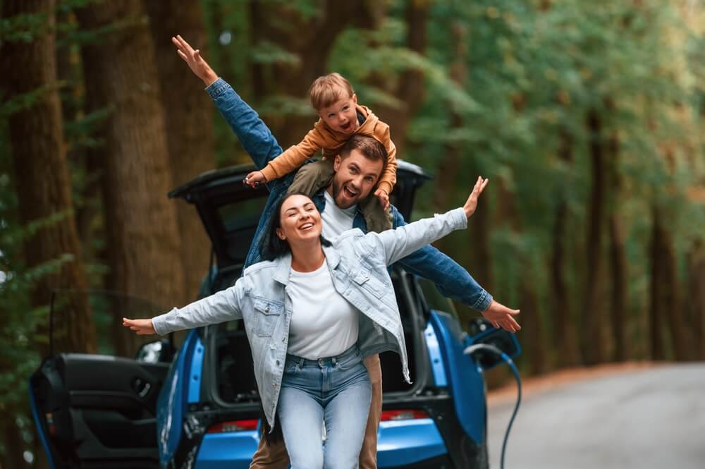 Happy, smiling family of man, woman and child act silly in the woods outside their car - best car insurance.