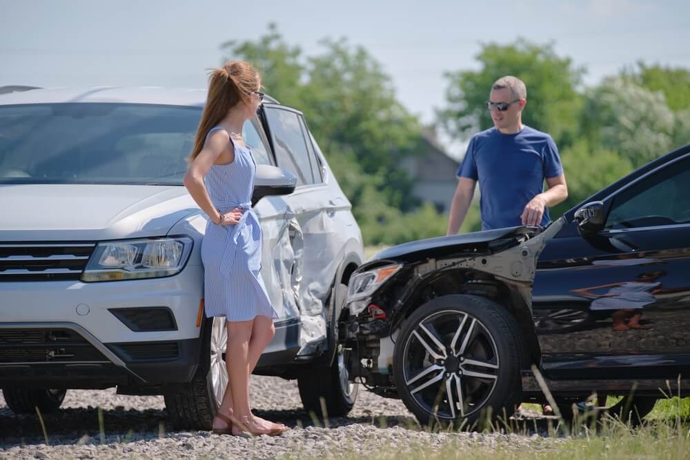Two drivers exchange comments after they get into a wreck, lady in blue dress, man in blue T-shirt, looking over hood of car at each other.