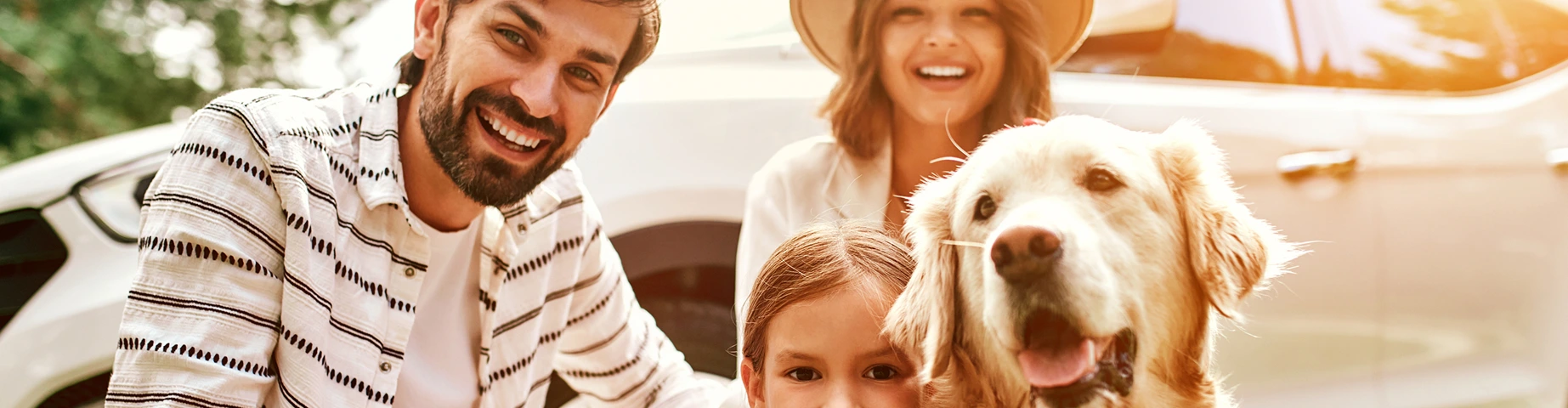 Family saves enough money by bundling with InsureOne to travel on a trip to nature for the weekend. Mom and Dad with their daughter and a Labrador dog are standing near the car.